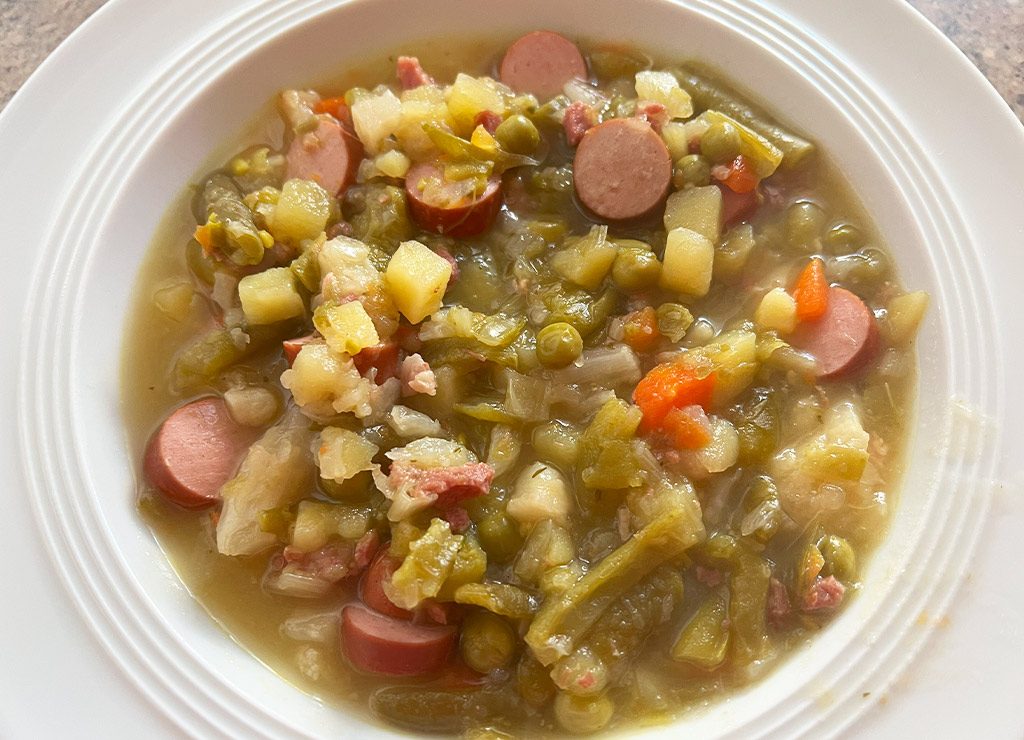 Nahaufnahme eines Tellers mit herzhaft dampfender Erbsensuppe, die mit Würstchen, Kartoffeln, Karotten und Erbsen gefüllt ist.