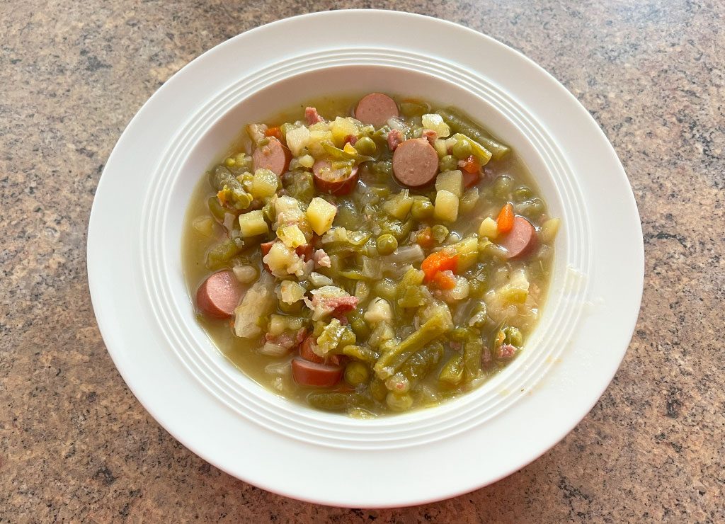 Ein Teller mit Erbsensuppe, serviert in einem weißen Porzellanteller auf einer gemusterten Tischoberfläche. Die Suppe enthält geschnittene Würstchen, Kartoffeln, Karotten und Erbsen.