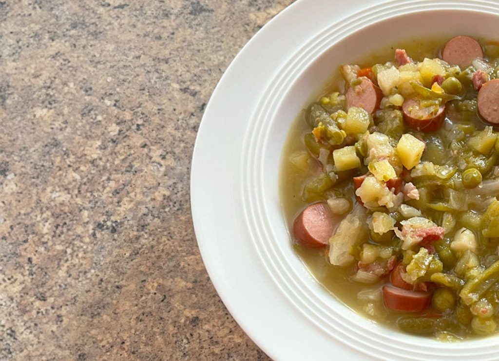 Teilansicht eines Tellers mit Erbsensuppe, platziert auf einer gemusterten Tischoberfläche. Die Suppe enthält Würstchen und Gemüse, darunter Karotten und Erbsen.