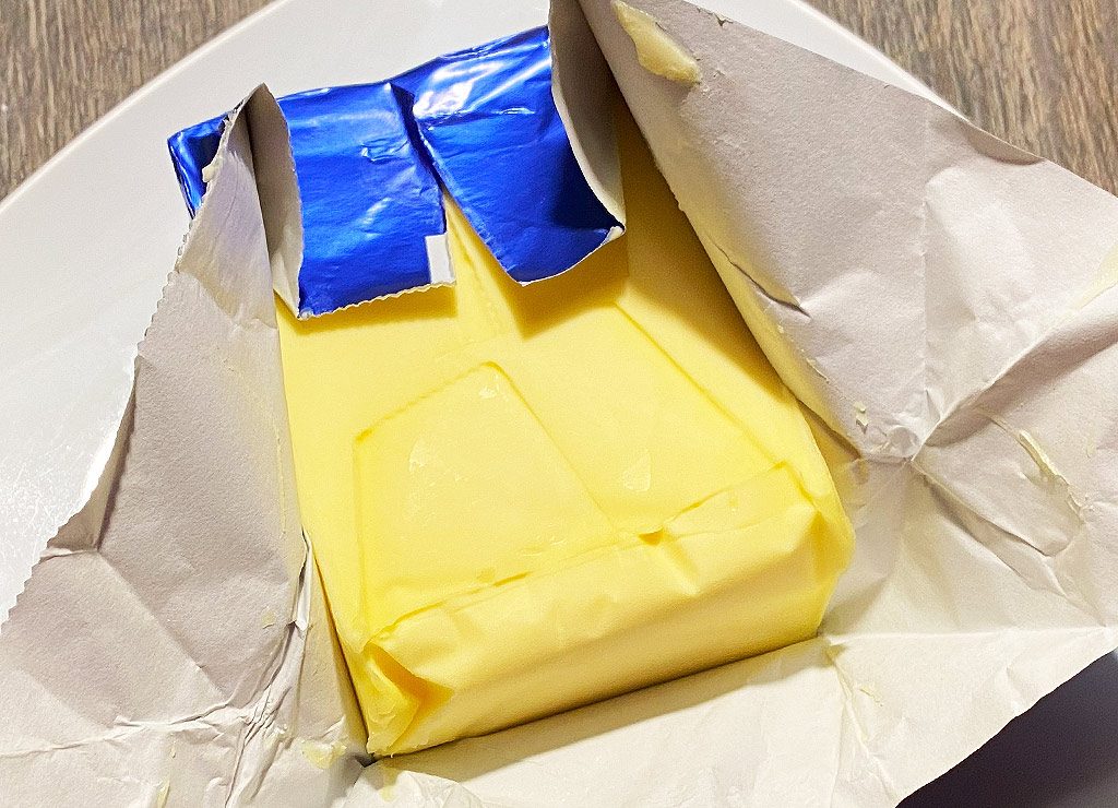 Geöffnete Butterverpackung mit gelbem Butterblock, die auf einem weißen Teller liegt.