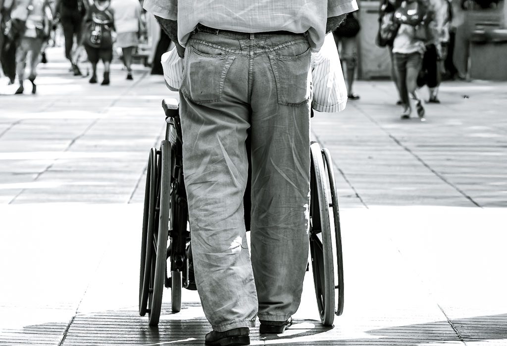 Tag der Tagespflegen in Deutschland. Kuriose Feiertage - 10. November © 2024 Sven Giese Mann schiebt eine Person im Rollstuhl auf einem belebten Gehweg, symbolisiert Unterstützung und Pflege.