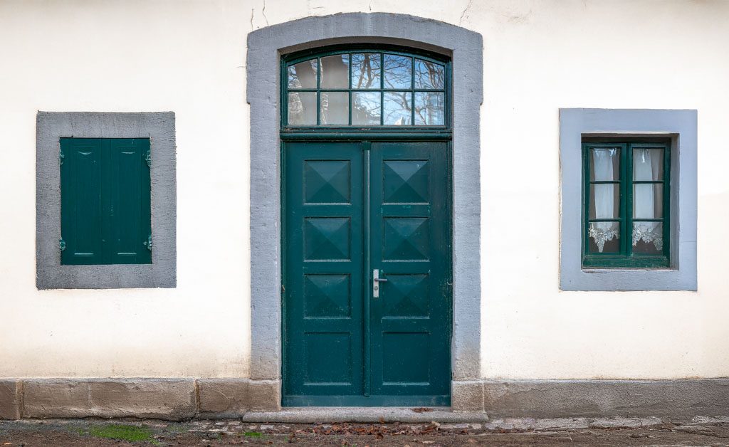 Hausfassade mit grüner Tür, Fensterläden links und Fenstern mit Vorhängen rechts.