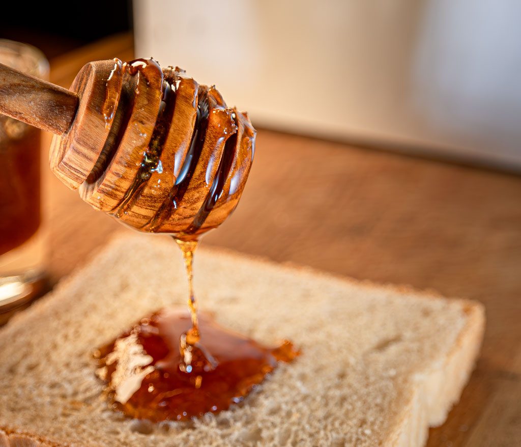 Holzhoniglöffel mit fließendem Honig über einer Scheibe Brot, im warmen Licht fotografiert.