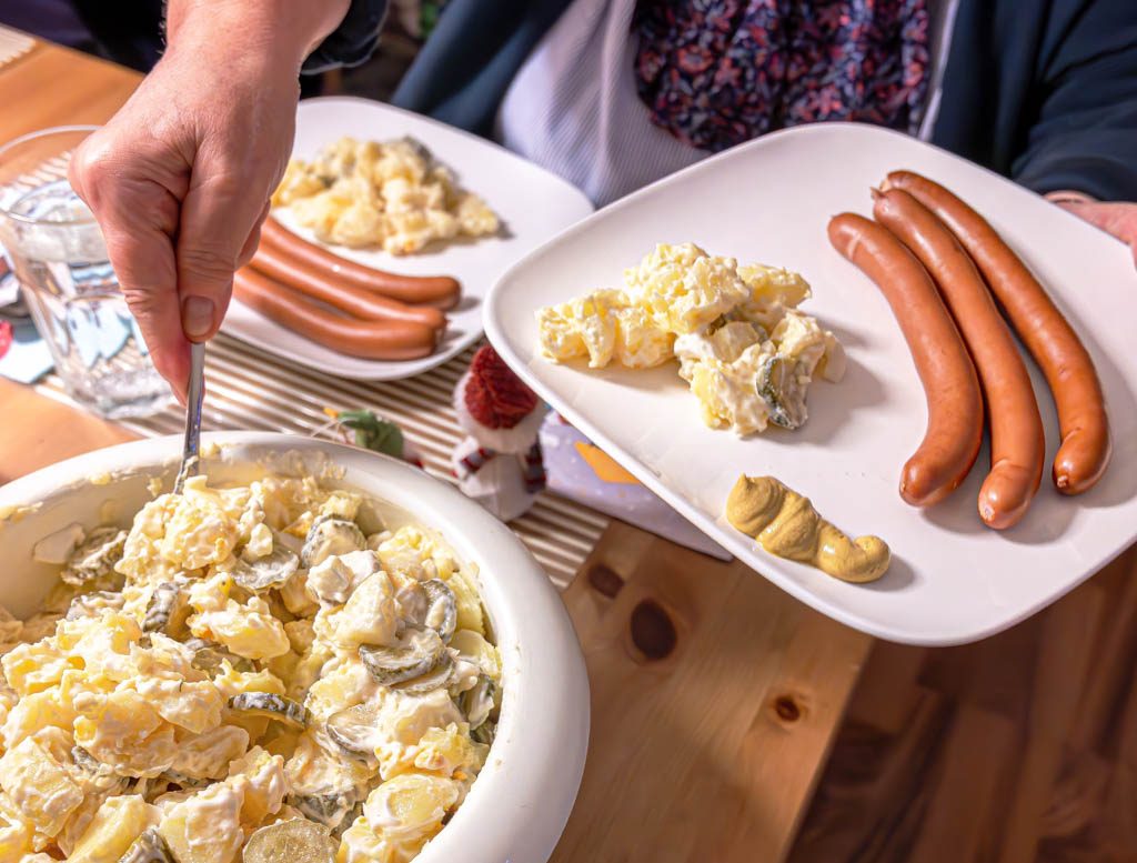 Eine Hand serviert Kartoffelsalat aus einer weißen Schüssel, daneben ein Teller mit Würstchen.