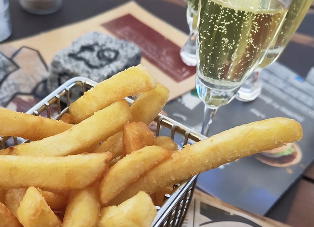 Eine silberne Frittierkorb-Schale mit Pommes frites neben einem gefüllten Champagnerglas auf einem Tisch.