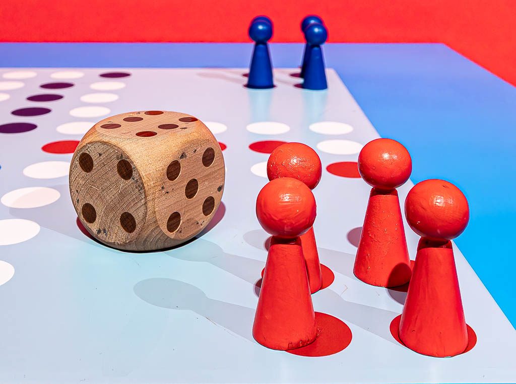 Rote und blaue Spielfiguren mit großem Holzwürfel auf buntem Spielbrett vor rotem Hintergrund.