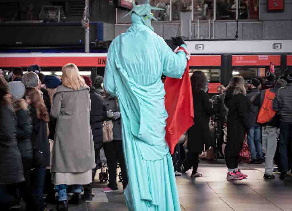 Sehr große Person im Kostüm der Freiheitsstatue steht mit rotem Tuch vor wartender Menschenmenge auf einem öffentlichen Platz.