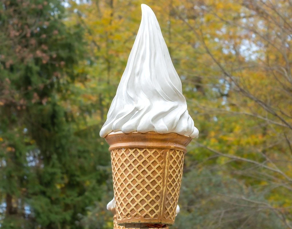 Große Softeis-Skulptur in Waffeloptik mit weißer Eisspitze an einem Verkaufsstand vor herbstlichen Bäumen.