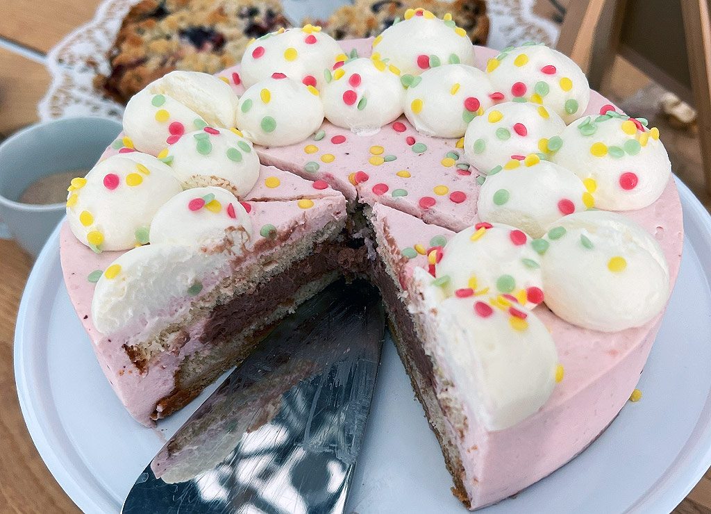 Tag der Eiscreme-Torte - National Ice Cream Pie Day in den USA. Kuriose Feiertage - 18. August (c) 2025 Sven Giese Rosa Eistorte mit Sahnetupfen und bunten Streuseln, angeschnitten auf einer weißen Tortenplatte.