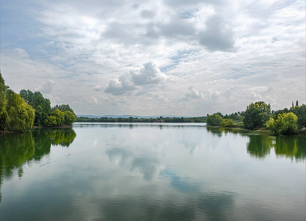 Welttag der Seen – UN World Lake Day. Kuriose Feiertage – 27. August (c) 2025 Sven Giese – Bild 2 Weitläufiger See mit Wolkenhimmel, umrahmt von grünen Bäumen und sanfter Landschaft.