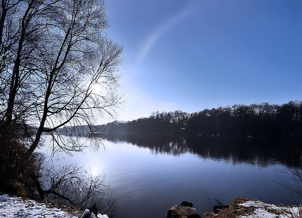 Welttag der Seen – UN World Lake Day. Kuriose Feiertage – 27. August (c) 2025 Sven Giese – Bild 3 Ruhiger See im Winter mit kahlen Bäumen, Sonnenreflexion und klarem blauen Himmel.
