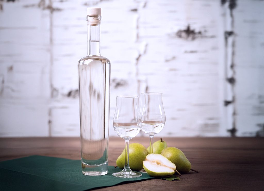 Klare Obstbrandflasche mit zwei Gläsern, frische Birnen daneben, halbierte Frucht vorn platziert, stehen auf einem Holztisch.