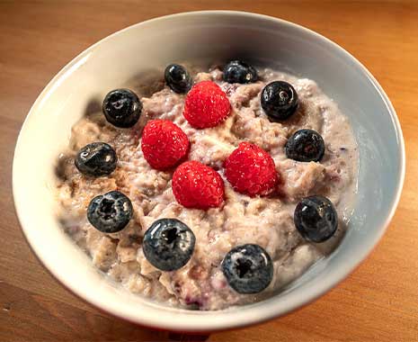 Weiße Schüssel Porridge mit Himbeeren und Blaubeeren, cremig, frisch serviert auf Holztisch.