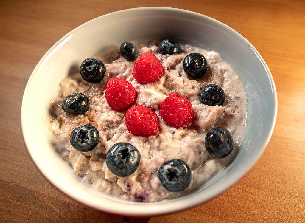 World Porridge Day – Welttag des Porridge. Kuriose Feiertage – 10. Oktober © 2025 Sven Giese - Bild 1 Weiße Schüssel Porridge mit Himbeeren und Blaubeeren, cremig, frisch serviert auf Holztisch.