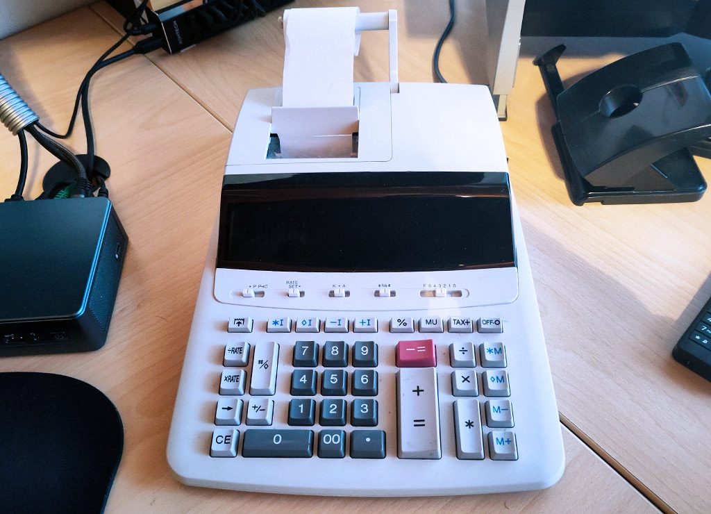 Großer Tischrechner mit Papierrolle steht auf Schreibtisch, umgeben von Kabeln und Geräten.