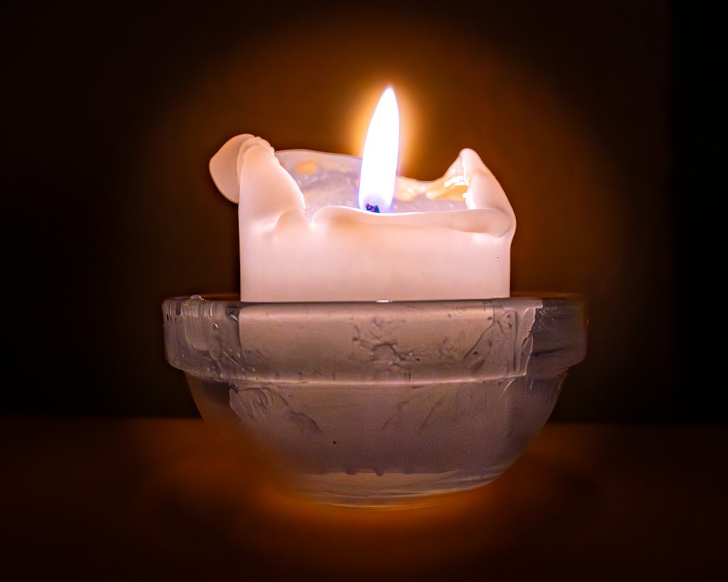 Brennende weiße Kerze in Glas-Schälchen, warmes Licht leuchtet sanft im dunklen Raum.