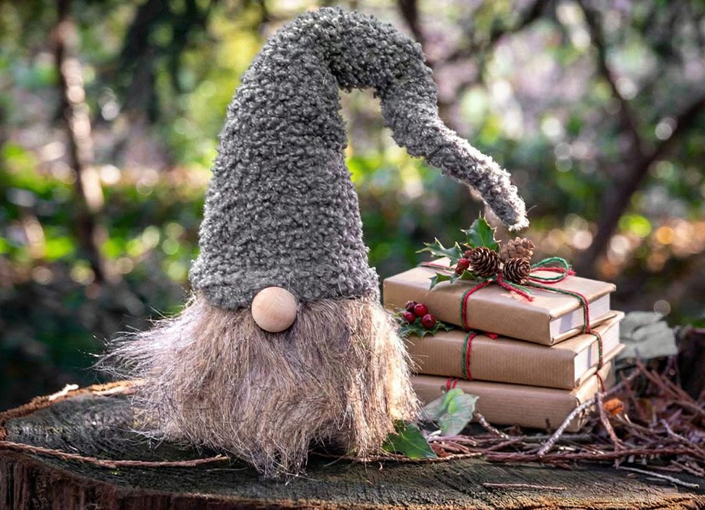 Gnomfigur mit grauer Filzmütze, daneben Bücherstapel mit Tannenzweig im Sonnenlicht auf Baumstumpf.