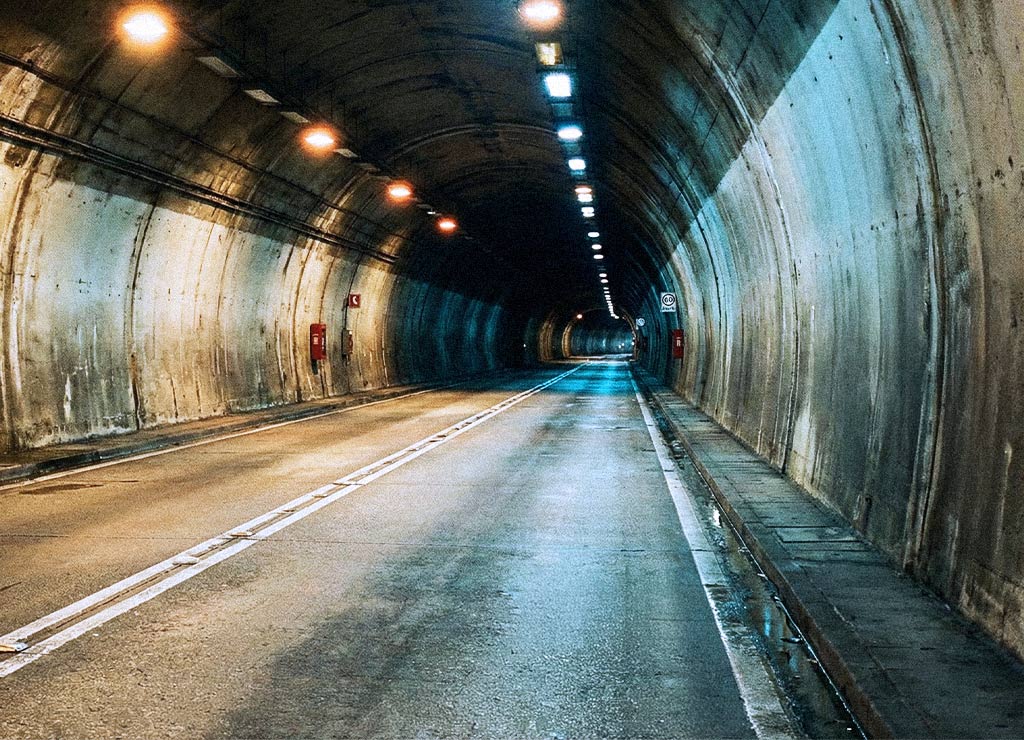 Leerer Straßentunnel mit Fahrbahn, gelber Deckenbeleuchtung, führt lange tief in die Dunkelheit.