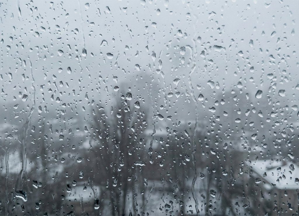 Nasse Fensterscheibe mit vielen Regentropfen, trüber Januartag mit verschwommenem Winterpanorama dahinter.