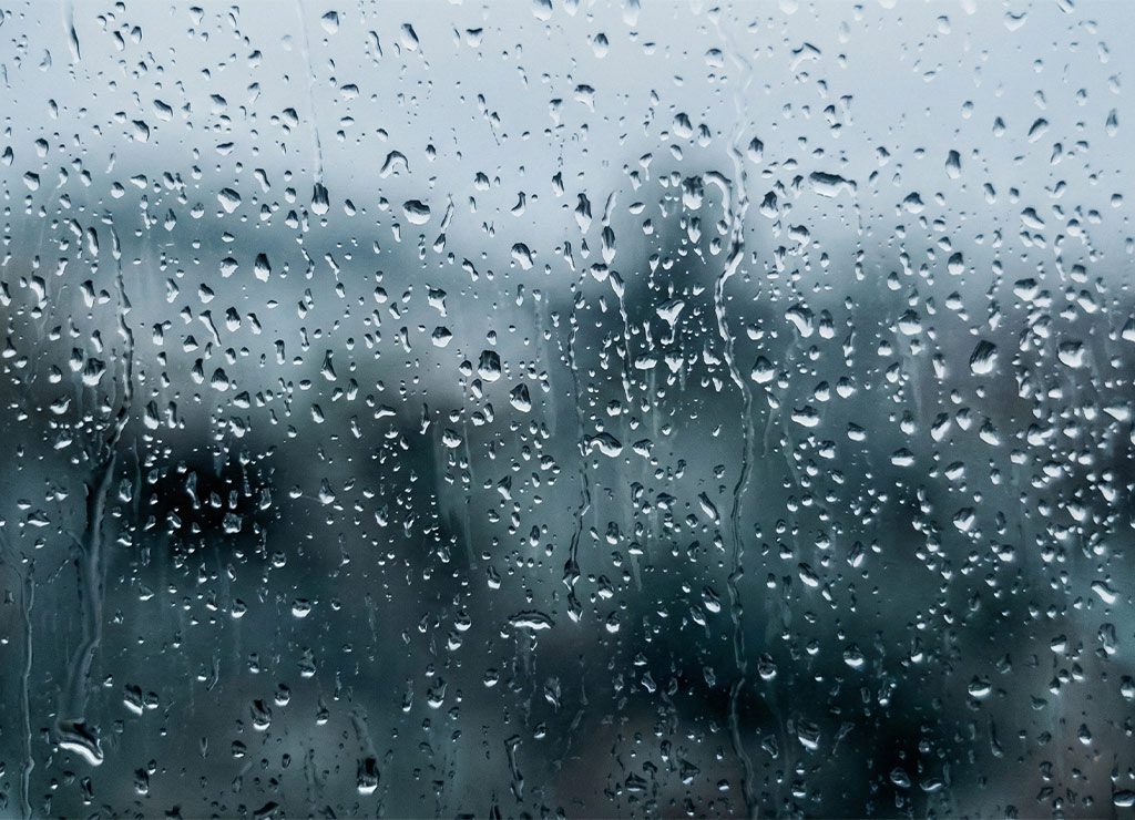 Regentropfen auf Fensterscheibe, grauer Winterhimmel und unscharfe Bäume oder Gebäude im Hintergrund, melancholische Stimmung.