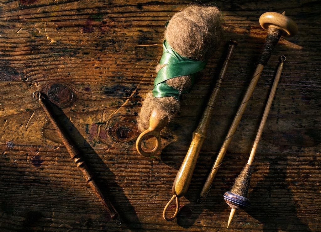 Spinnwerkzeuge und Wollbüschel locker arrangiert auf altem Holztisch im warmen Seitenlicht.