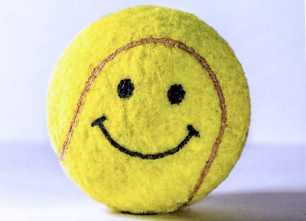 Gelber Tennisball mit aufgemaltem Smiley liegt auf Tisch vor hellem, weißem Hintergrund.