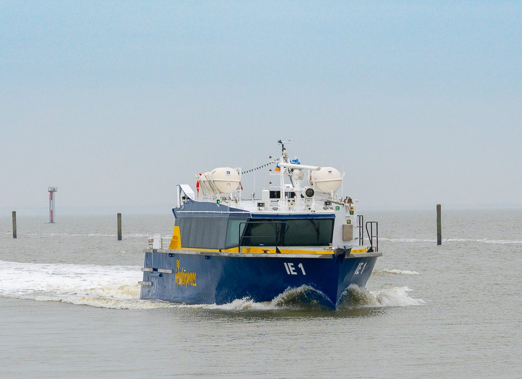 Tag der Fähren. Kuriose Feiertage – 22. Februar (c) 2026 Sven Giese – Bild 2 Blaues Schiff „IE 1“ fährt durch graues Wattenmeer, Bugwelle spritzt, Pfähle im Hintergrund.