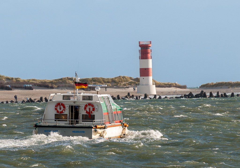 Tag der Fähren. Kuriose Feiertage – 22. Februar (c) 2026 Sven Giese – Bild 3 Kleine Fähre auf dem Weg zur Düne Helgoland fährt durch wellige Nordsee, im Hintergrund Leuchtturm und Dünen am Strand.