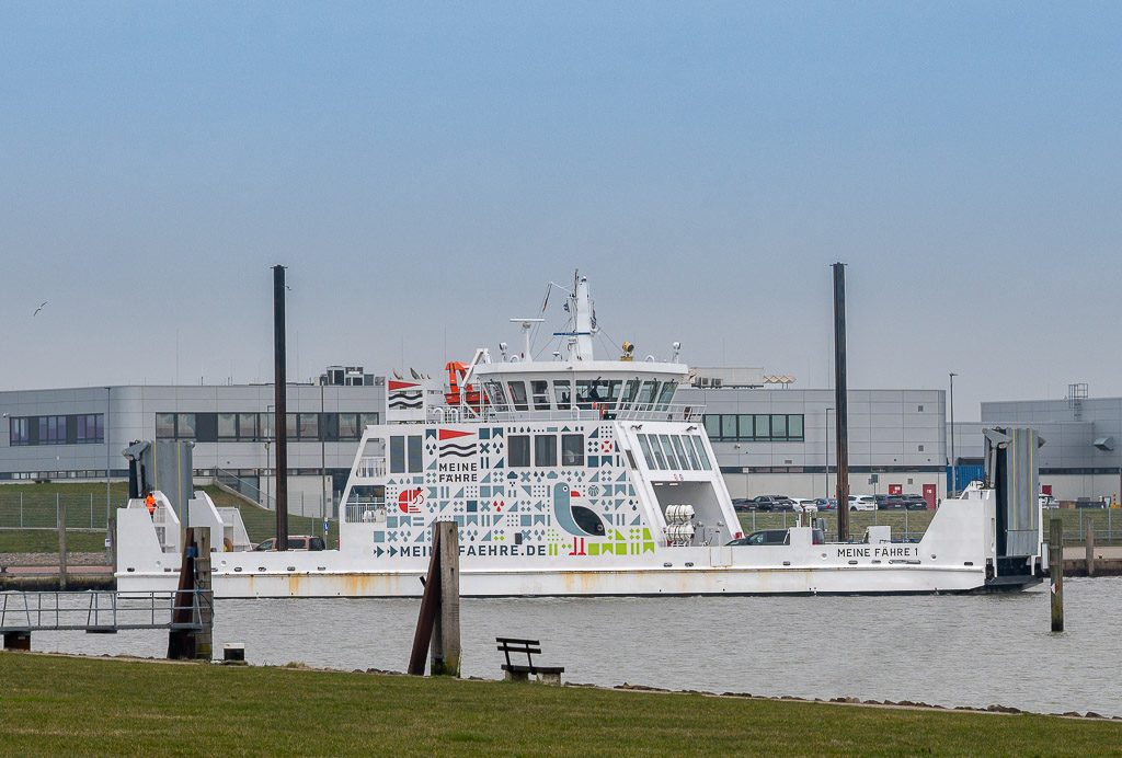 Tag der Fähren. Kuriose Feiertage – 22. Februar (c) 2026 Sven Giese – Bild 1 Weiße Autofähre „Meine Fähre 1“ bei der Einfahrt in den Hafen, davor Wiese mit einer Bank und Industriegebäuden im Hintergrund.
