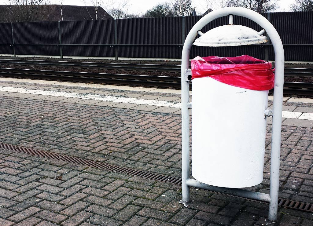 International Day of Zero Waste Day. Kuriose Feiertage - 30. März (c) 2026 Sven Giese - Bild 2 Weißer Mülleimer mit rotem Müllsack steht am Bahnsteig vor Gleisen und Zaun.
