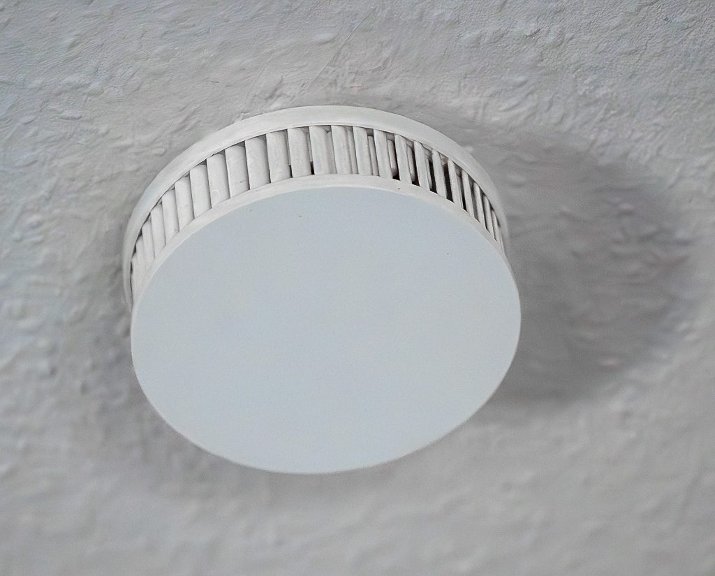 Weißer Rauchmelder an rauer Decke, schräg fotografiert, Lüftungsschlitze im Wohnraum klar sichtbar.