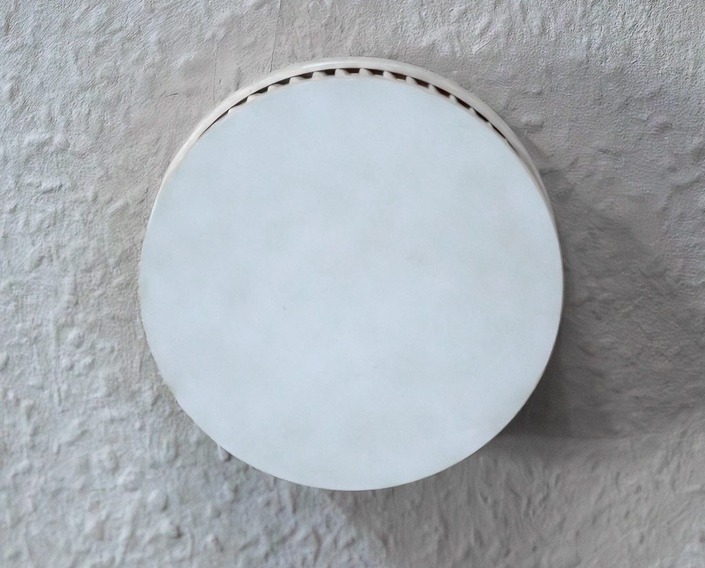 Weißer Rauchmelder an rauer Decke, frontal von unten aufgenommen, runde Form zentral im Bild.