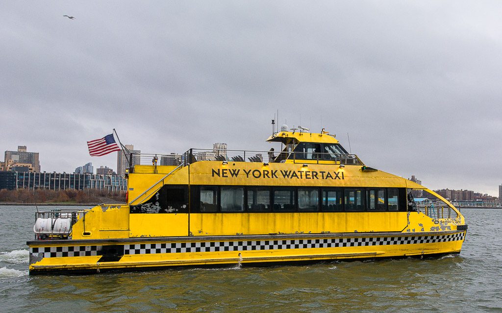 Gelbes New York Wassertaxi mit US-Flagge fährt nah am Ufer über graues Wasser.