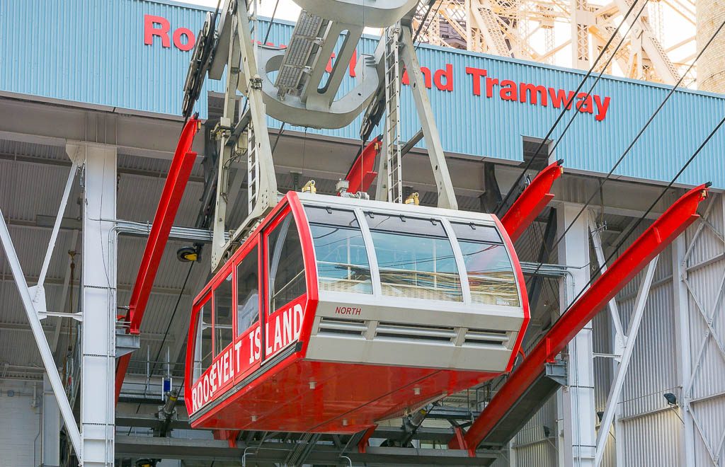 Rote Roosevelt-Island-Seilbahn hängt hoch oben an Stahlkonstruktion vor Stationsgebäude in New York.
