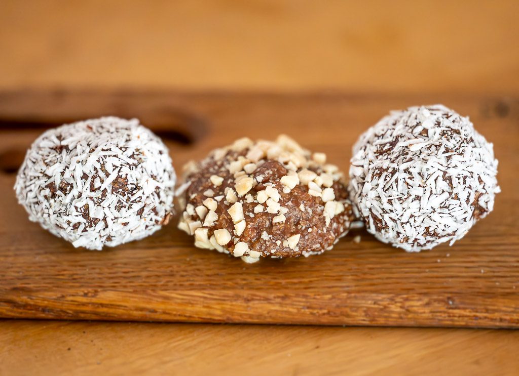 Drei Schokoladenbällchen mit Kokos und Nüssen liegen auf Holzbrett vor hellem Hintergrund.