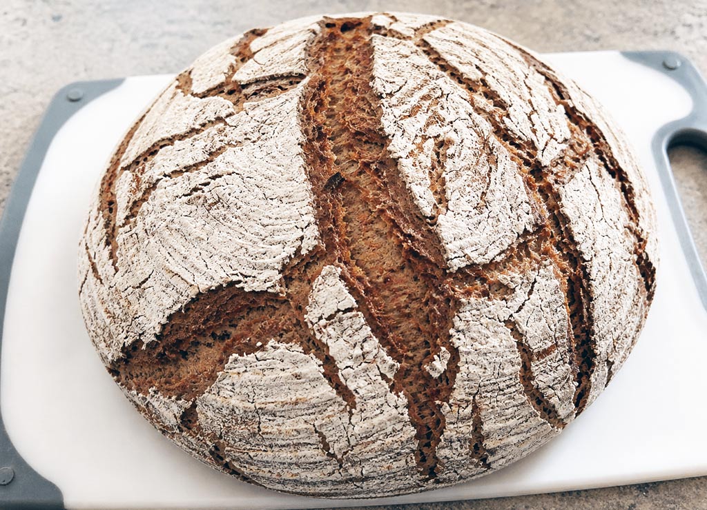 Runder Brotlaib mit rustikaler Kruste und Mehlrissen liegt auf weißem Schneidebrett in Küche.