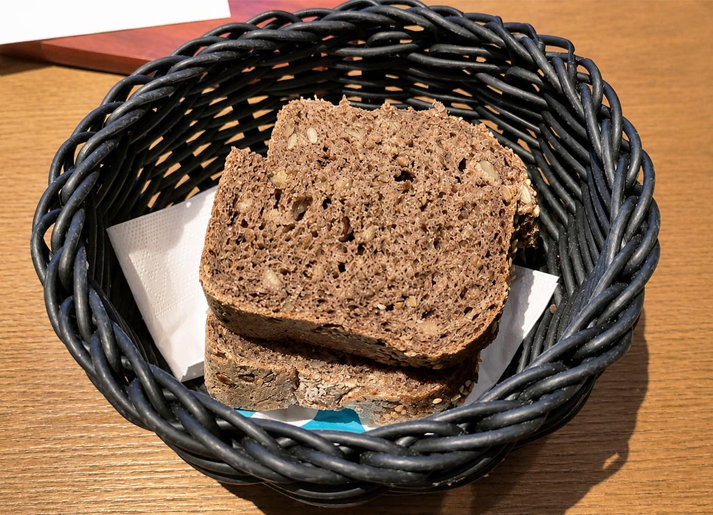 Dunkle Brotscheiben liegen in schwarzem Brotkorb auf Holztisch, Körner im Anschnitt sichtbar.