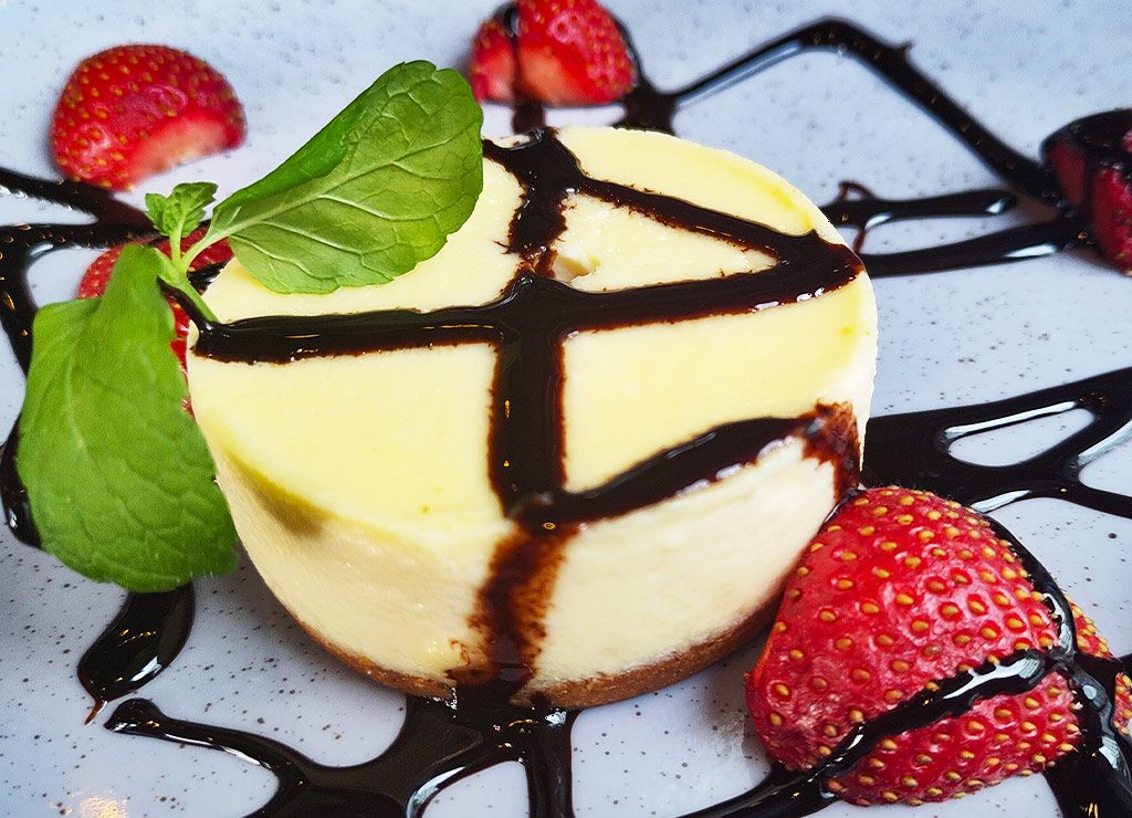 Tag des Erdbeerkäsekuchens – Strawberry Cheesecake Day. Kuriose Feiertage – 8. Juni (c) 2026 Sven Giese – Bild 3 Kleiner Strawberry Cheesecake mit Schokosauce, Erdbeeren und Minze auf weißem Teller.