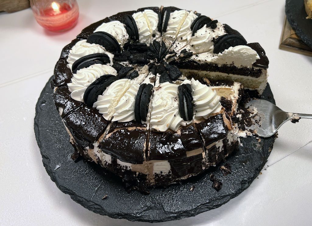 International Cake Day – Internationaler Tag des Kuchens. Kuriose Feiertage – 20. Juli (c) 2026 Sven Giese – Bild 1 Angeschnittener Schokoladenkuchen mit Sahne, dunklen Keksen und Glasur liegt auf schwarzer Platte.