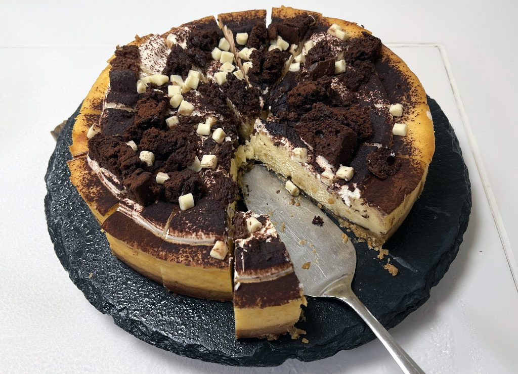 International Cake Day – Internationaler Tag des Kuchens. Kuriose Feiertage – 20. Juli (c) 2026 Sven Giese – Bild 2 Käsekuchen mit Brownies, Kakaopulver und weißer Schokolade ist auf dunkler Platte angeschnitten.