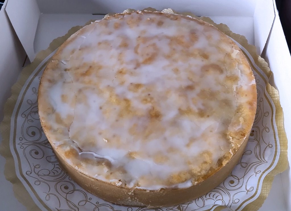 International Cake Day – Internationaler Tag des Kuchens. Kuriose Feiertage – 20. Juli (c) 2026 Sven Giese – Bild 3 Runder Apfelkuchen mit hellem Zuckerguss liegt auf einer verzierten Pappunterlage im Karton.