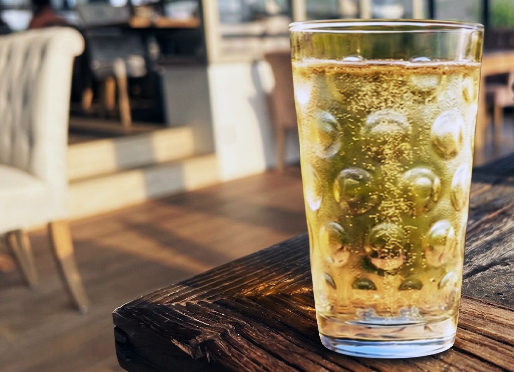 Tag der Schorle in Deutschland. Kuriose Feiertage – 23. Juli (c) 2026 Sven Giese – Bild 1 Weinschorle im hohen Dubbeglas mit Noppen steht auf rustikalem Holztisch im Sonnenlicht.