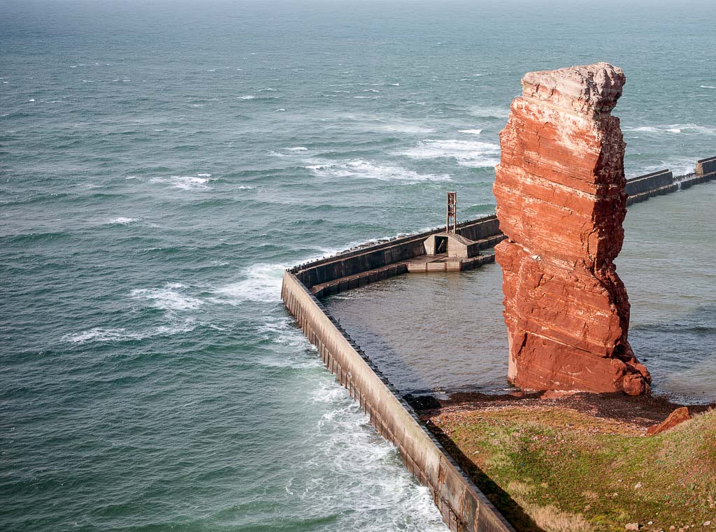 Der rote Felsmonolith Lange Anna ragt an Helgolands Küste neben Brandung und Schutzmauern empor.
