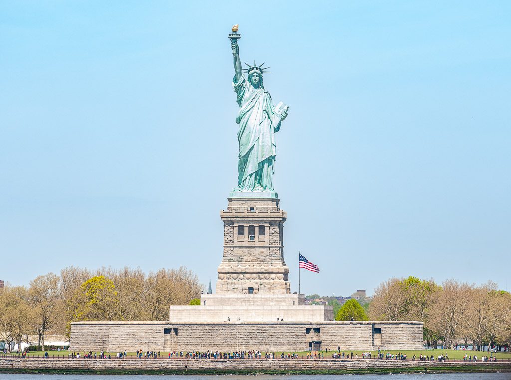 Geburtstag der Freiheitsstatue in New York – Statue of Liberty Dedication Day USA Kuriose Feiertage – 28. Oktober © 2026 Sven Giese – Bild 1 Die Freiheitsstatue auf Liberty Island mit amerikanischer Flagge, Bäumen und Besuchern am Ufer.