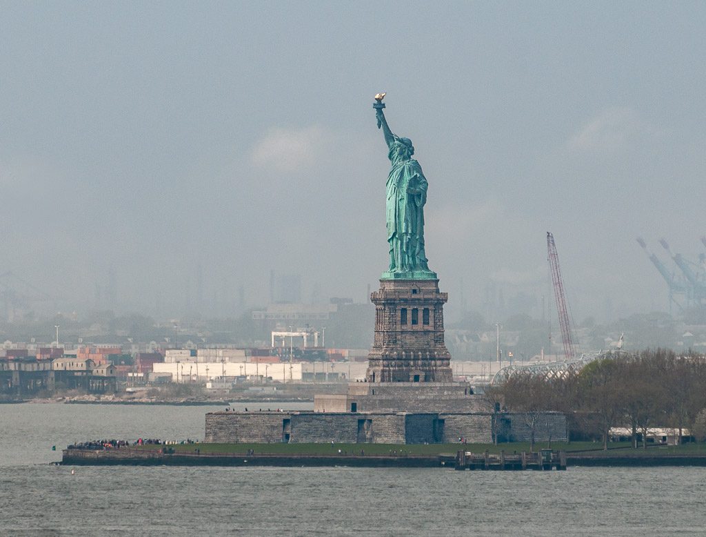 Geburtstag der Freiheitsstatue in New York – Statue of Liberty Dedication Day USA Kuriose Feiertage – 28. Oktober © 2026 Sven Giese – Bild 2 Die Freiheitsstatue steht auf Liberty Island vor diesigem Himmel, vom Wasser aus fotografiert.