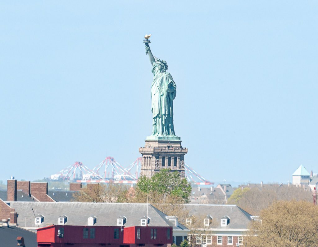 Geburtstag der Freiheitsstatue in New York – Statue of Liberty Dedication Day USA Kuriose Feiertage – 28. Oktober © 2026 Sven Giese – Bild 4 Seitliche Ansicht der Freiheitsstatue über New Yorker Dächern, Hafenkräne stehen im Hintergrund.