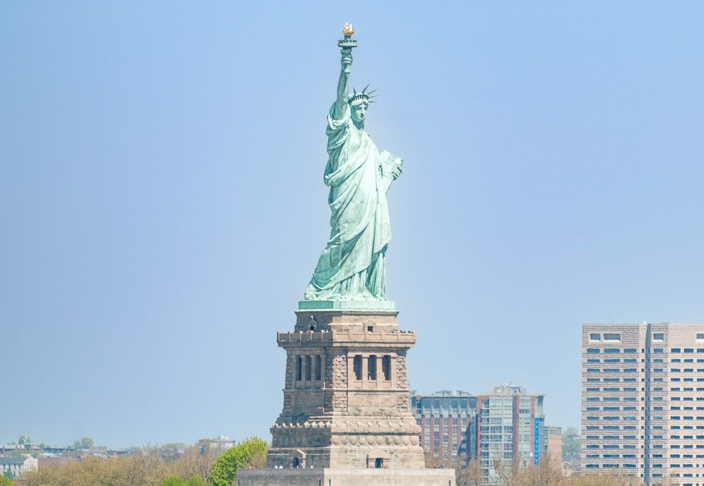 Geburtstag der Freiheitsstatue in New York – Statue of Liberty Dedication Day USA Kuriose Feiertage – 28. Oktober © 2026 Sven Giese – Bild 5 Die Freiheitsstatue ragt vor blauem Himmel auf, rechts stehen hohe Gebäude der Stadt.