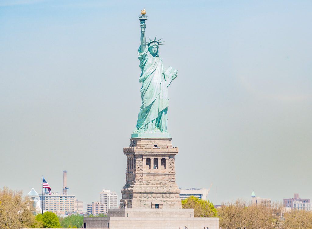 Geburtstag der Freiheitsstatue in New York – Statue of Liberty Dedication Day USA Kuriose Feiertage – 28. Oktober © 2026 Sven Giese – Bild 7 Freiheitsstatue auf Liberty Island mit erhobener Fackel, dahinter New Yorker Gebäude und US-Flagge.